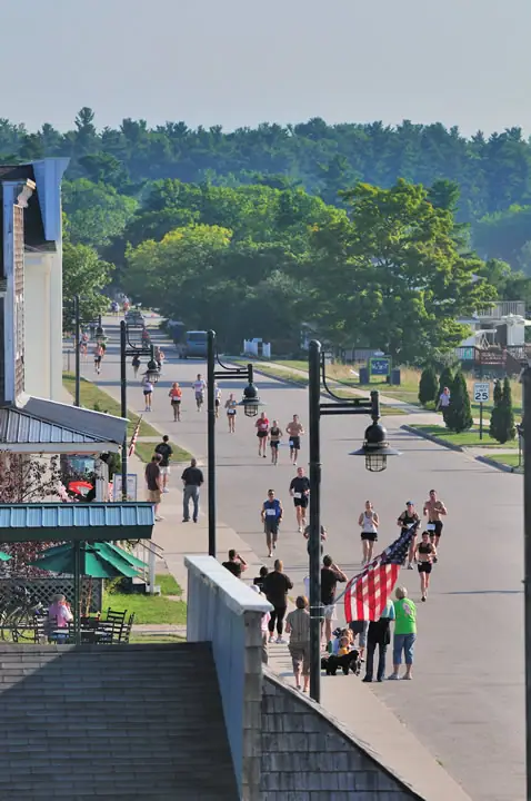 Runners move down a street as people carry a large American flag and spectators watch from the sidewalk.
