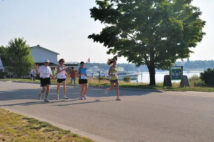 Four runners move along a waterfront road beside a dock, trees, buildings, and boats.