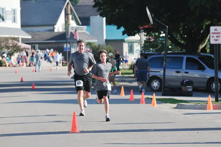 Two runners move down a cone-lined street while spectators gather near houses and parked vehicles.