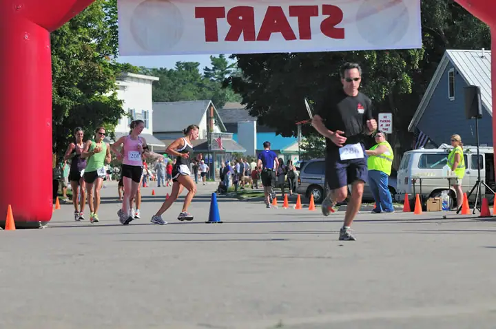 Runners pass beneath a red inflatable arch while spectators and volunteers line a street.