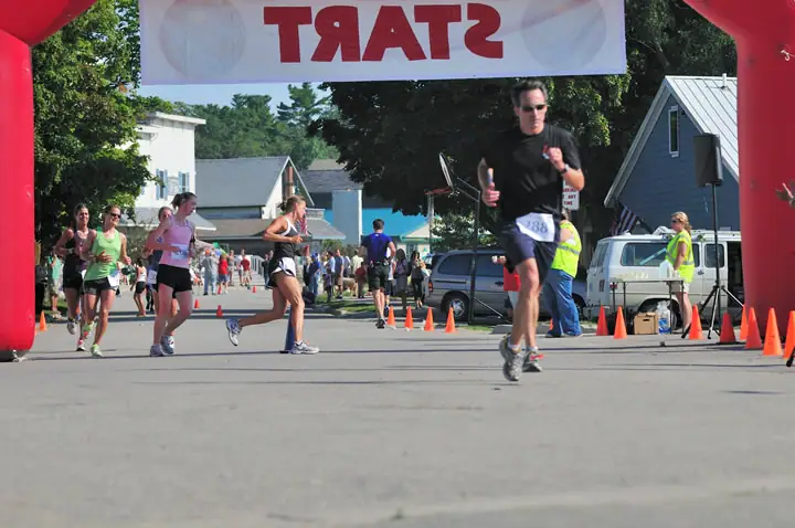 Runners pass beneath a red inflatable arch on a street lined with traffic cones and spectators.