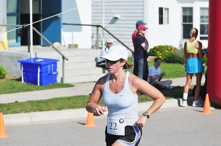A woman runner wearing bib number 22 passes traffic cones as spectators gather outside a building.