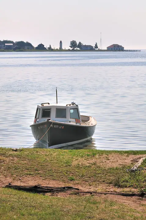 A cabin boat is moored at a grassy shoreline, with a lighthouse, buildings, antenna, and pier across the water.