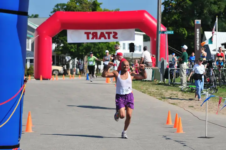 A runner raises his arms while approaching a red inflatable arch on a cone-marked street.