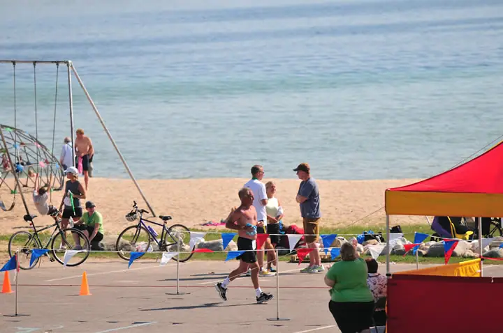 A runner passes a beachside course marked with flags and cones as spectators gather near bicycles and a bright tent.