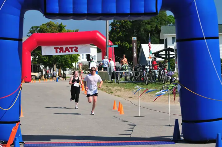 Two runners approach a blue inflatable race arch while spectators and bicycles line a paved street.