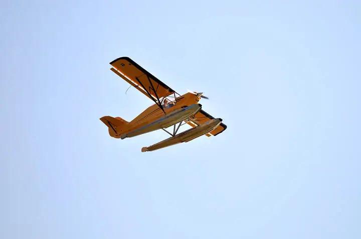 A yellow biplane with twin floats flies against a pale blue sky.