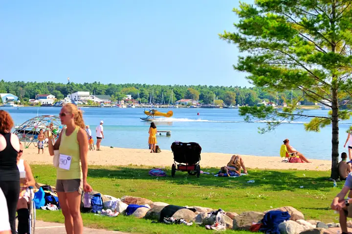 People gather on a sandy beach beside blue water as a yellow seaplane passes offshore.