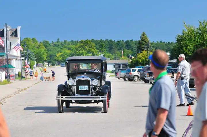 A black vintage automobile approaches on a paved street as spectators watch from the roadside.