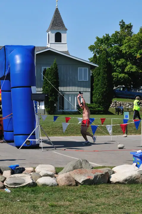 A shirtless athlete throws a shot put beside a blue inflatable structure, with a steepled church behind him.