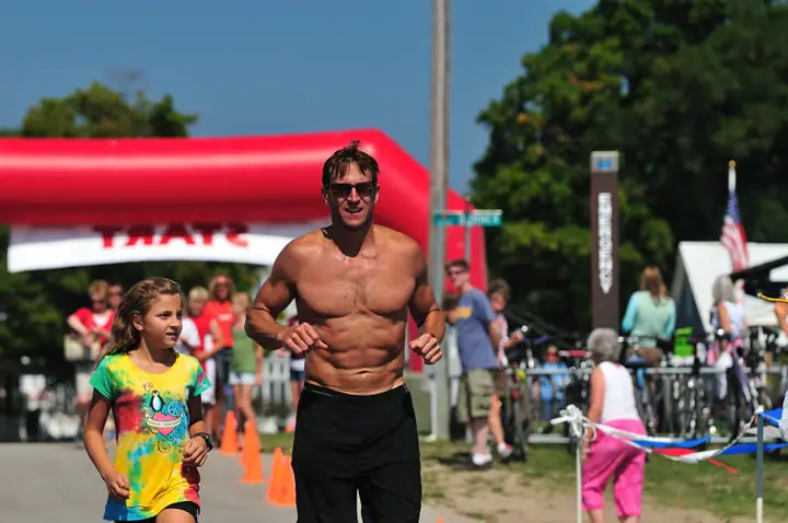 An adult runner and a child run toward the camera at an outdoor race finish area.