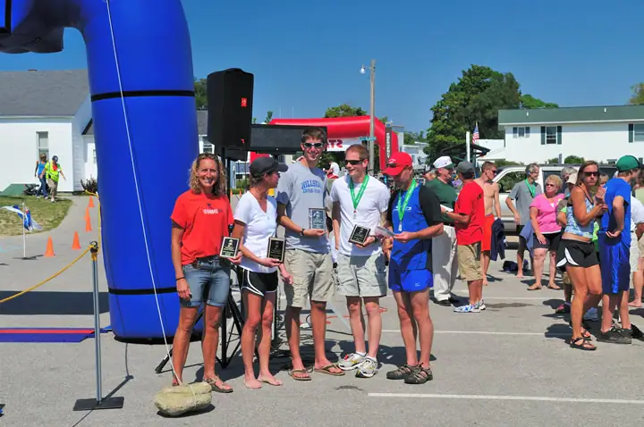 Five people display plaques and medals beside a blue inflatable arch while spectators gather behind them.