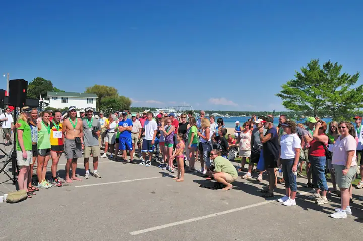 A large crowd of runners and spectators gathers on a paved waterfront area beneath a clear blue sky.