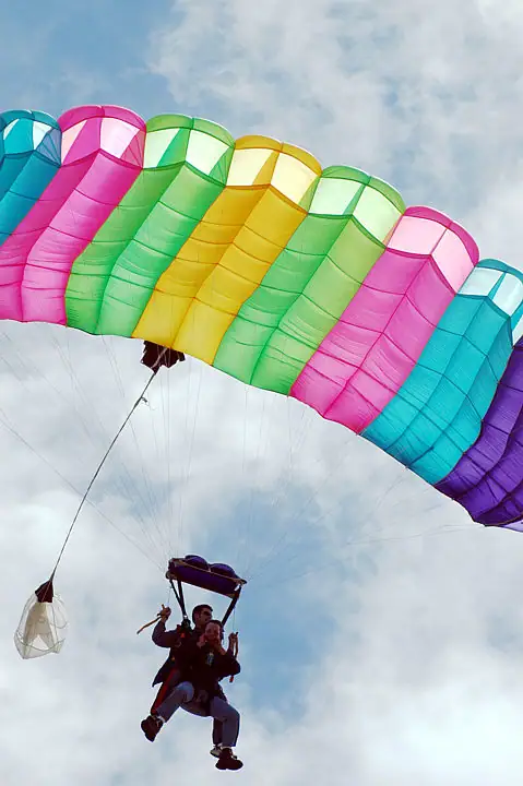 Two skydivers hang beneath a brightly striped parachute against a cloudy sky.
