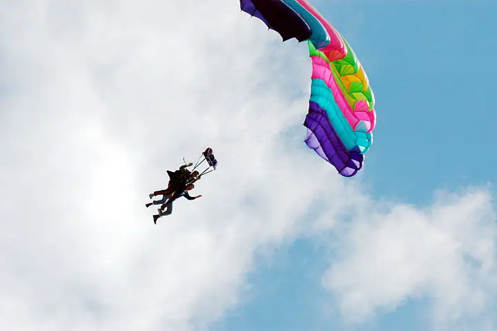Two skydivers descend beneath a brightly colored parachute against a blue, cloud-filled sky.