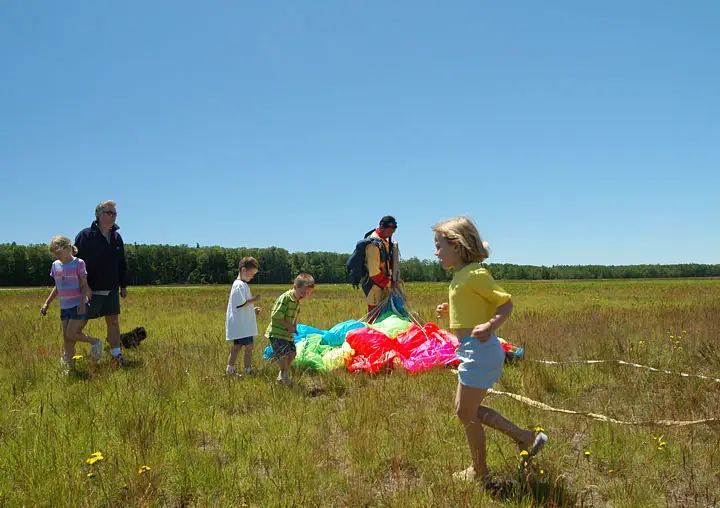 Adults and children gather around a colorful parachute in an open grassy field.