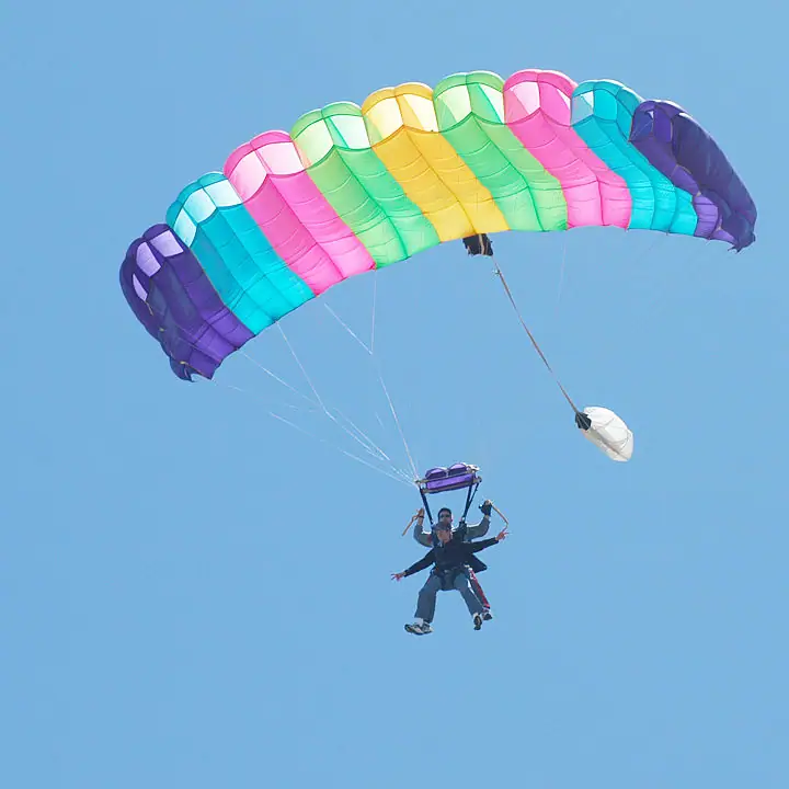 Two people descend beneath a brightly striped parachute against a clear blue sky.