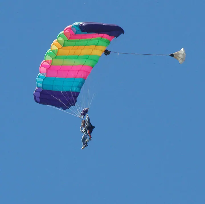 Two skydivers descend beneath a brightly striped parachute against a clear blue sky.