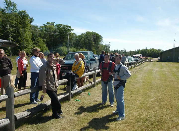 People gather beside a wooden fence while two wear parachute harnesses in a grassy field.