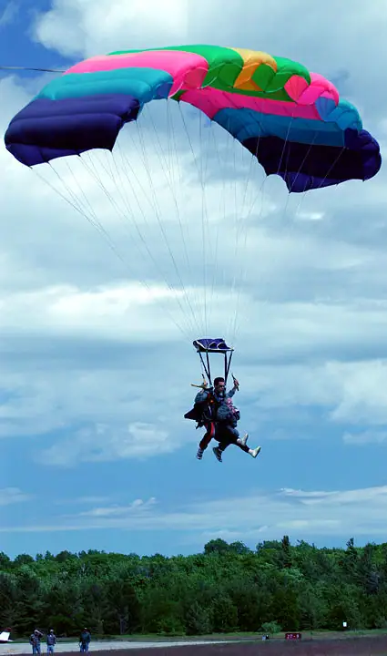 Two people descend beneath a brightly colored parachute over a wooded area and paved ground.