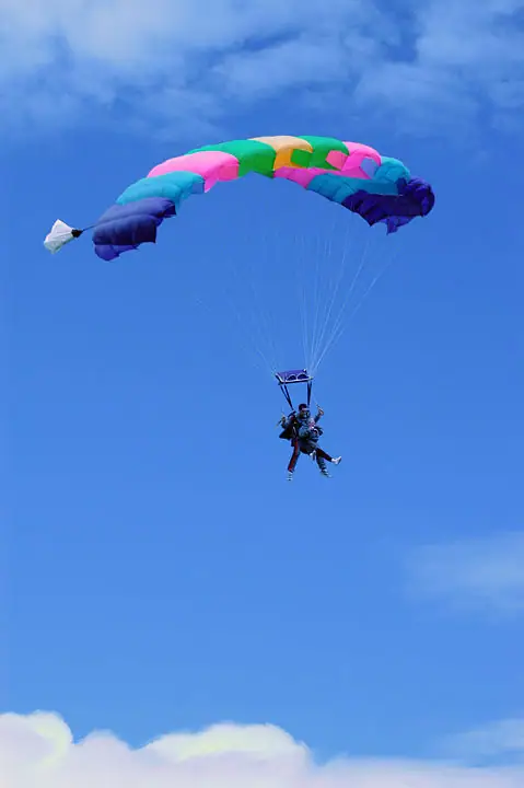Two people descend beneath a bright multicolored parachute against a blue, cloud-filled sky.