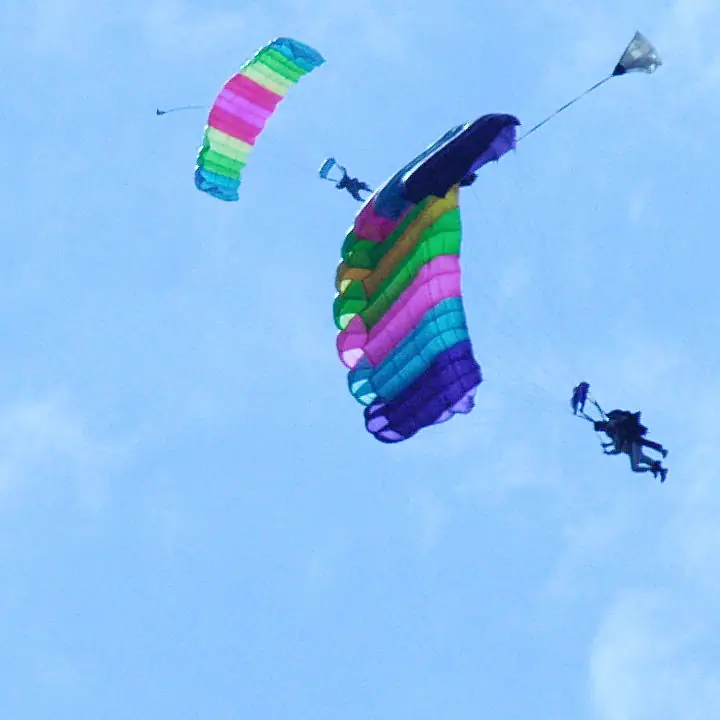 Two skydivers descend beneath brightly striped parachutes against a pale blue sky.