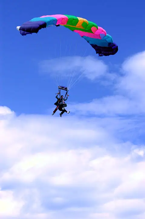 Two skydivers descend beneath a brightly colored parachute against a blue, cloud-filled sky.