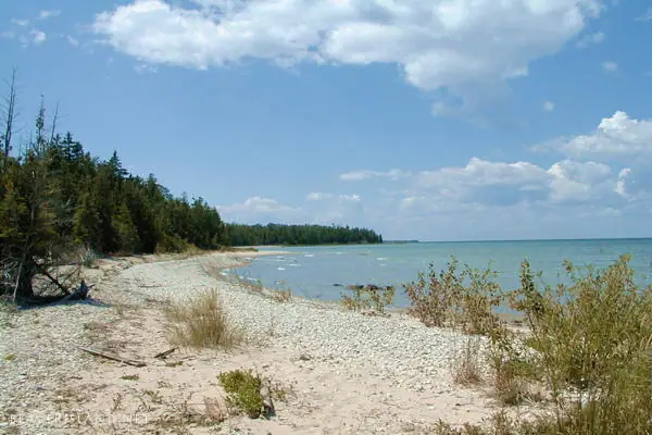 A broad pebble shoreline curves beside calm blue water, with forest along the left side under a partly cloudy sky.