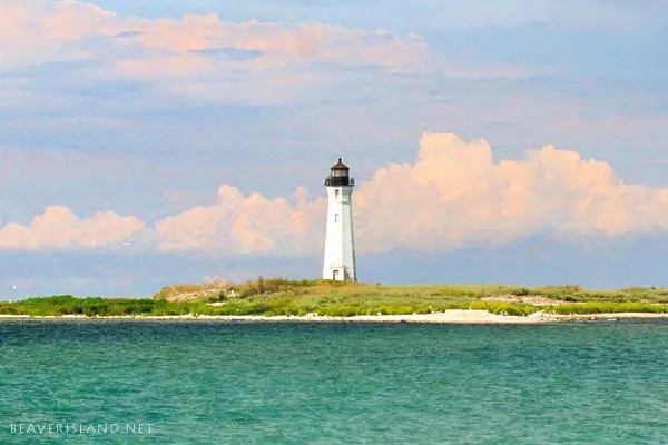White lighthouse on a low, green shoreline beyond blue-green water.