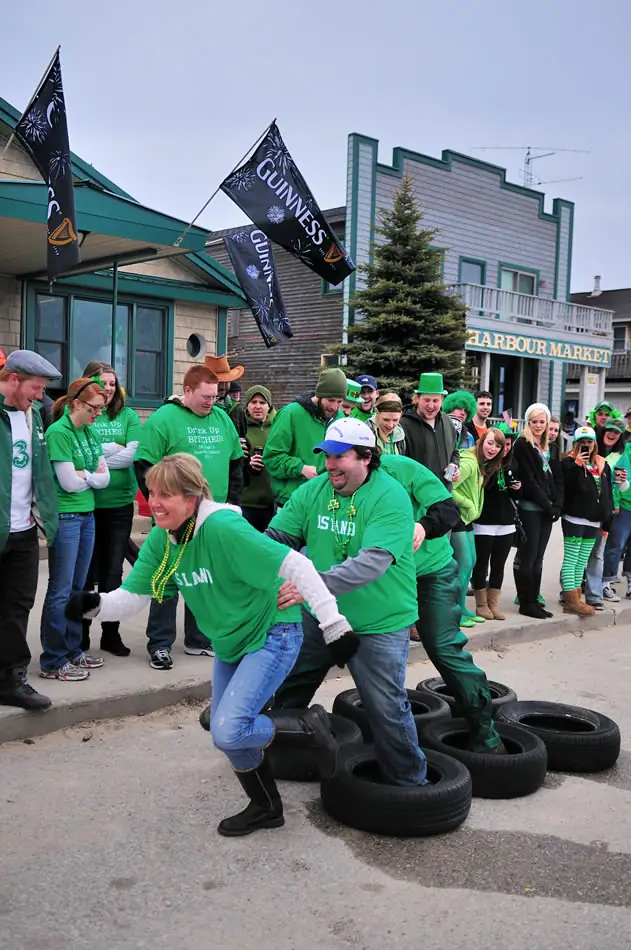 People in green clothing take part in a tire game beside a storefront as a crowd watches.
