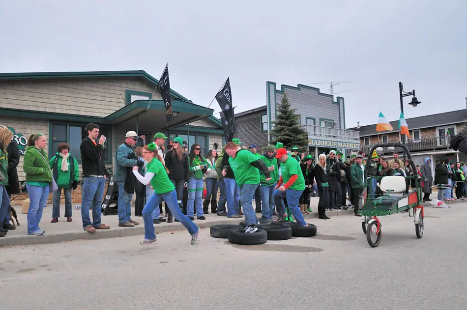 People in green clothing take part in a tire obstacle activity on a street beside a storefront and watching crowd.