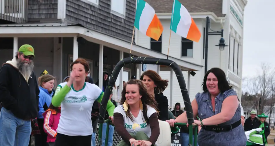 People pull a decorated parade vehicle beneath green-white-orange flags on a street lined with buildings.