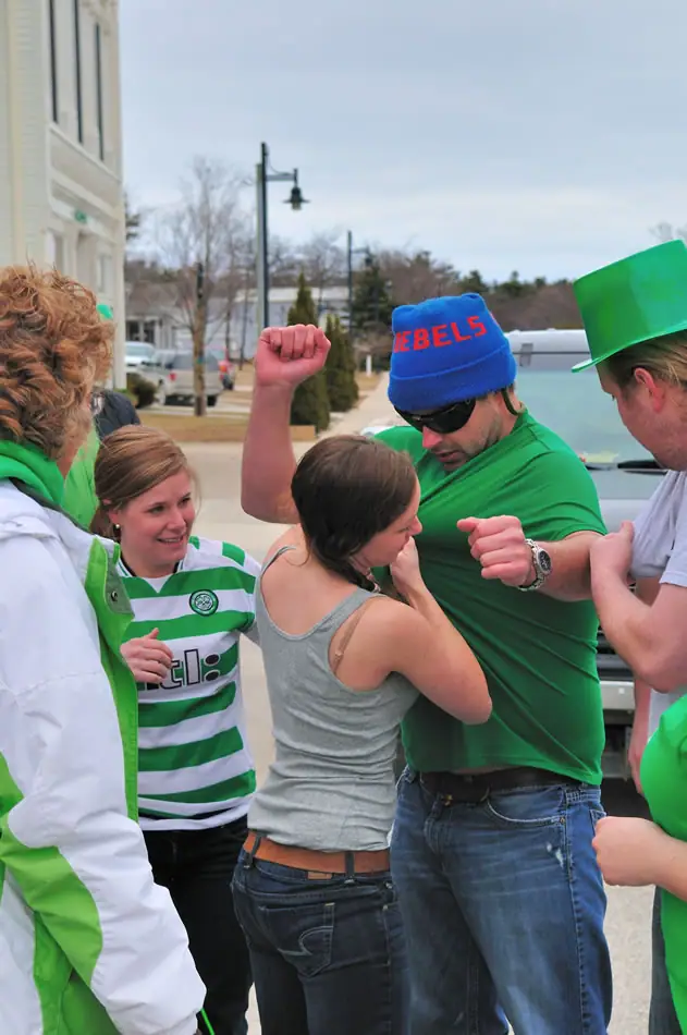 Adults in green clothing gather on a street, with one person raising a fist and wearing a blue REBELS beanie.