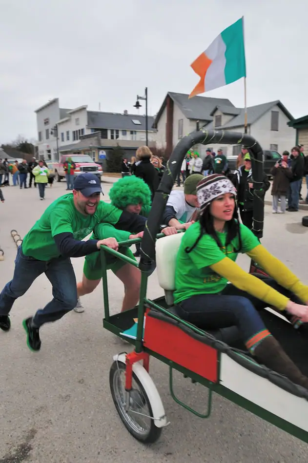 People in green clothing ride and push a decorated pedal-powered cart along a street beneath a tricolor flag.