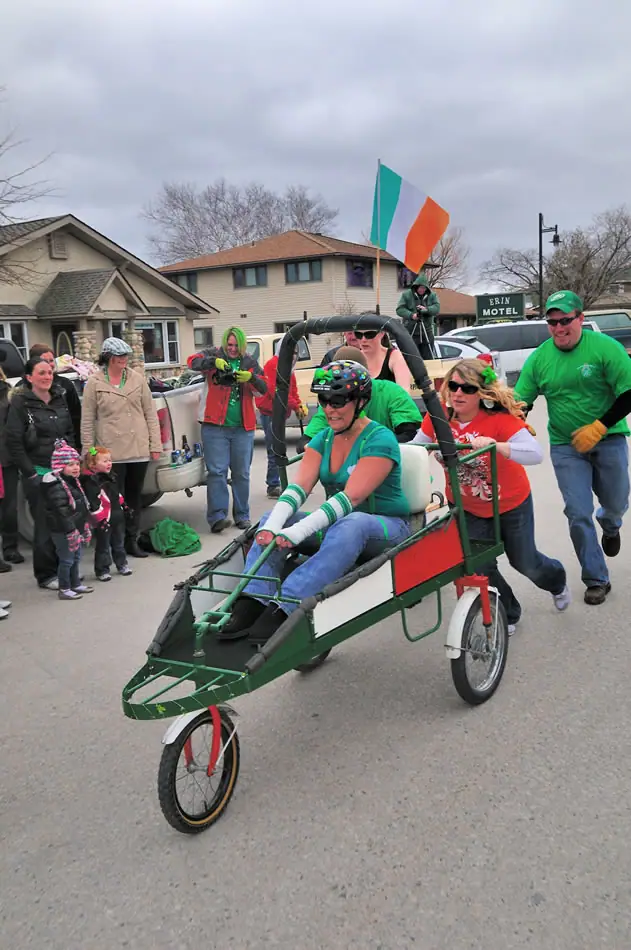 People ride and push a decorated green three-wheeled vehicle past spectators on a street.