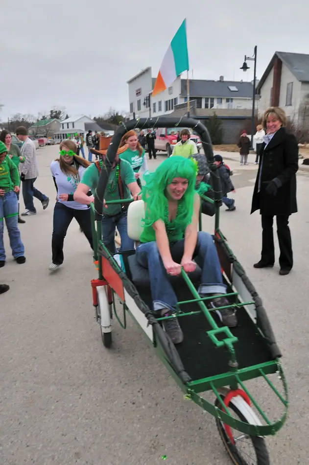 People in green gather around a large pedal-powered vehicle on a street during a festive procession.