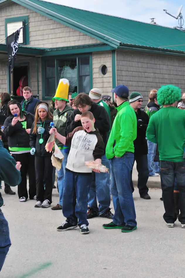 A crowd in green clothing gathers outside a shingled building, including a child in a sweatshirt and adults in novelty hats.