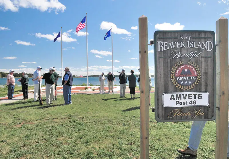 A wooden veterans’ sign stands beside three flagpoles and a group of people near the water.