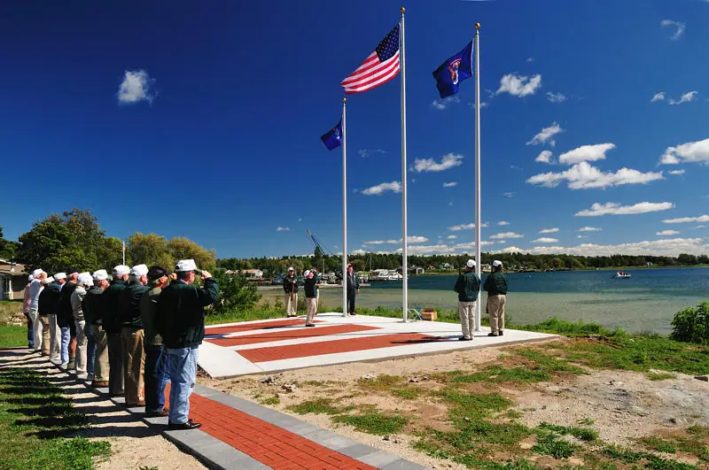 People salute beside three flagpoles at a waterfront ceremony.