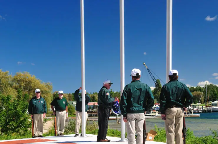 People in matching green jackets gather around three flagpoles above a marina and boats.