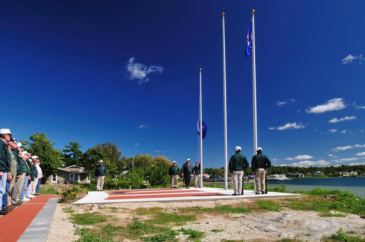 People gather beside flagpoles at a waterfront memorial with flags, a paved walkway, houses, and boats in the background.