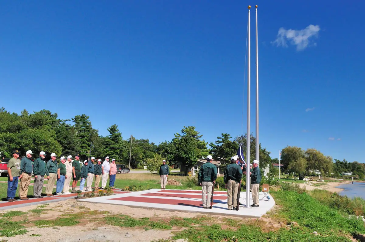 People gather beside striped flagpoles during an outdoor flag-raising ceremony near the water.