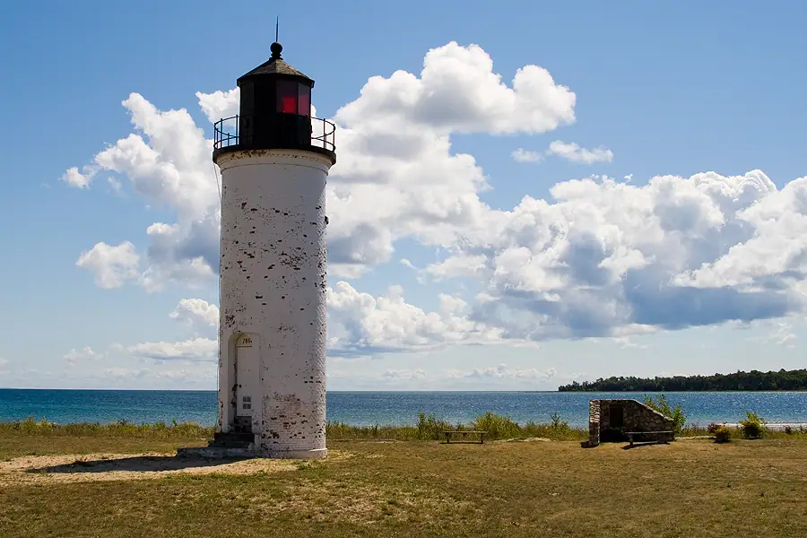 White cylindrical lighthouse beside a wooded shoreline under a cloud-filled blue sky.