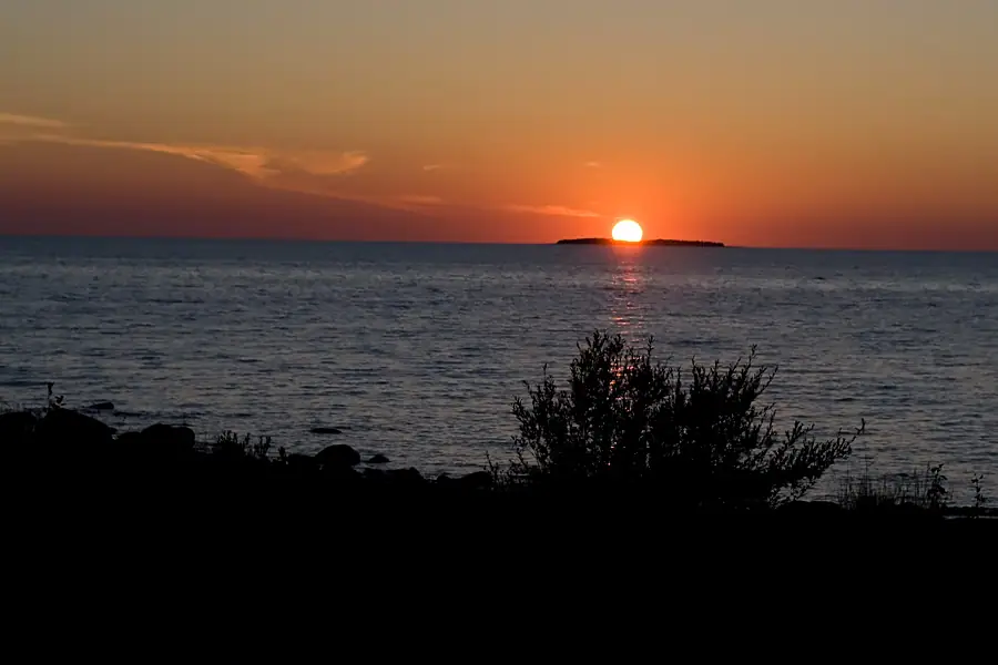 Sunset over rippling water, with a low dark landform on the horizon and silhouetted rocks and shrubs in the foreground.