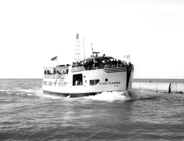 A crowded ferry moves beside a concrete breakwater, with passengers on its decks and flags flying.