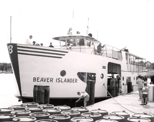 Black-and-white photograph of a white ferry at a pier with people and rows of wooden barrels.