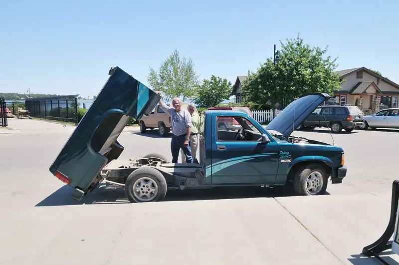 Two adults stand beside a blue pickup truck with its hood and rear body raised.