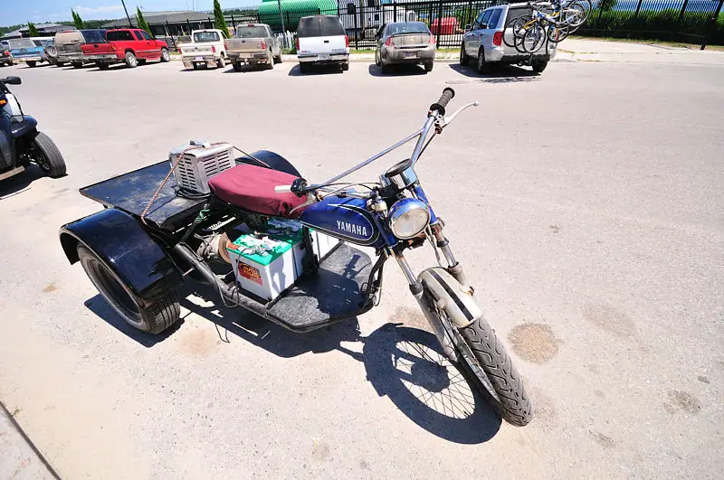 A blue Yamaha three-wheeled motorcycle with a cargo platform is parked in a paved lot among cars.
