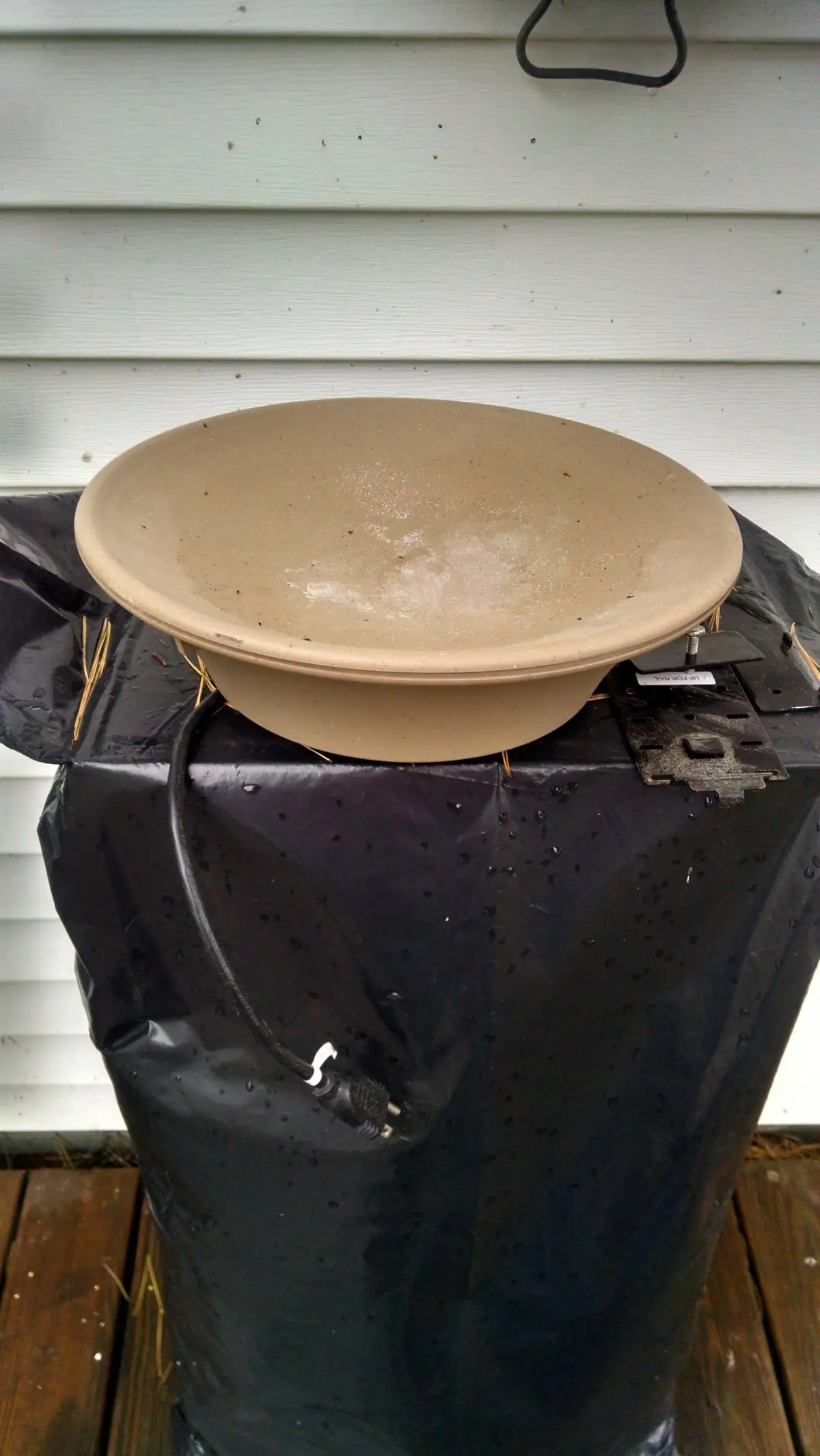 A shallow tan birdbath rests on a black plastic-covered container beside white horizontal siding.