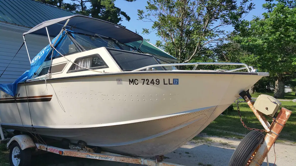 A white cabin motorboat rests on a trailer beside a building and trees.
